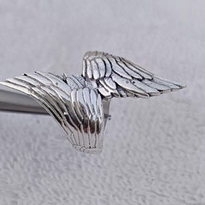 May include: A silver-colored ring featuring a detailed wing design. The ring's wings are spread out, with each feather individually sculpted. The ring is shown against a neutral background, highlighting the intricate details of the jewelry.
