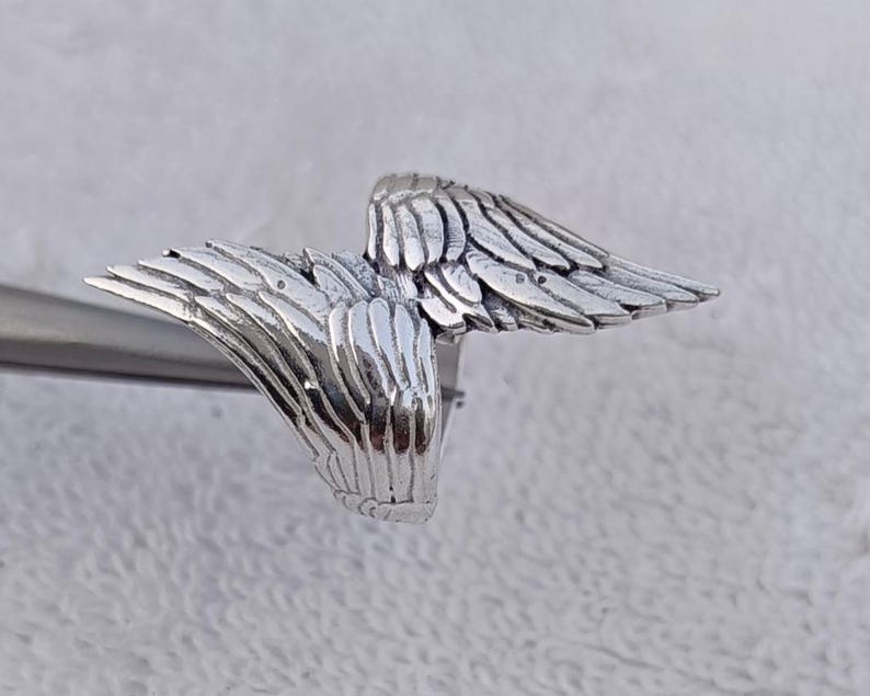 May include: A silver-colored ring featuring a detailed wing design. The ring's wings are spread out, with each feather individually sculpted. The ring is shown against a neutral background, highlighting the intricate details of the jewelry.