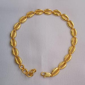 May include: A gold-toned bracelet composed of a series of interconnected cowrie shell-shaped links. The chain features a lobster claw clasp at one end and a short extender chain with circular links, creating a continuous circular arrangement.