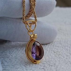 May include: A gold-toned pendant necklace featuring a pear-shaped, faceted purple amethyst gemstone set in a decorative metal frame with a twisted rope border. The pendant hangs from a matching gold-toned twisted chain link necklace.