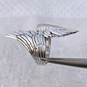 May include: A silver-colored ring with a detailed wing design. The wings are spread out on either side of the ring's band, with each feather individually sculpted. The ring is held by a pair of tweezers.