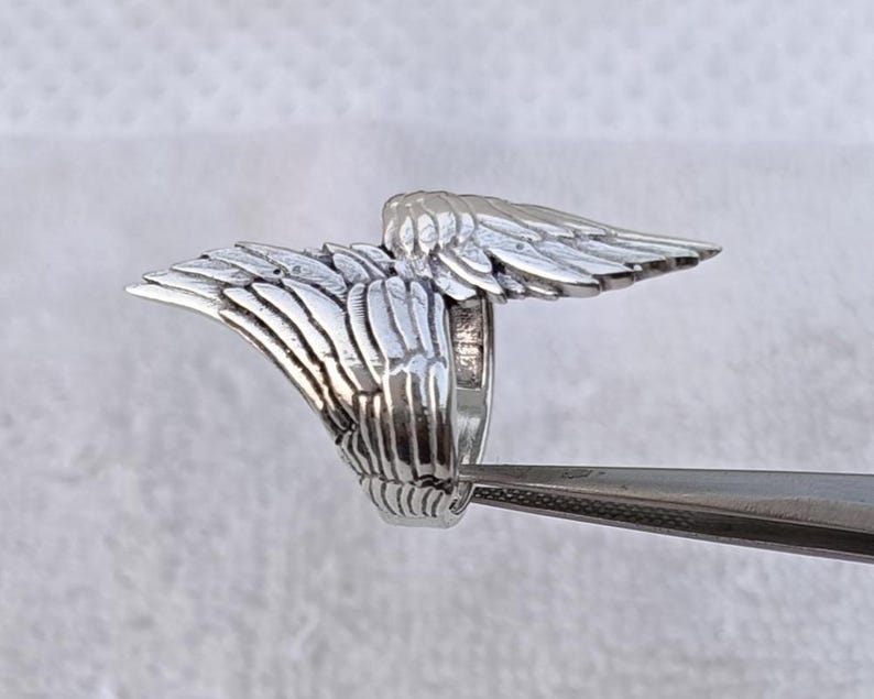 May include: A silver-colored ring with a detailed wing design. The wings are spread out on either side of the ring's band, with each feather individually sculpted. The ring is held by a pair of tweezers.