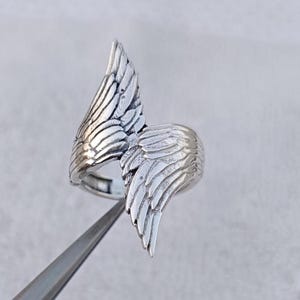 May include: A silver-colored ring featuring a detailed wing design. The ring has two wings that curve around the finger, with intricate feather detailing. The ring is held by a pair of tweezers.