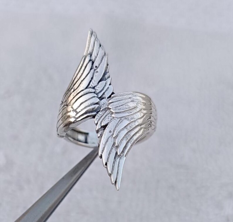May include: A silver-colored ring featuring a detailed wing design. The ring has two wings that curve around the finger, with intricate feather detailing. The ring is held by a pair of tweezers.