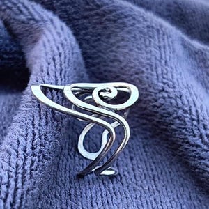 May include: A silver-colored ring with an abstract design. The ring features a swirling, openwork pattern with curved lines and a central spiral. The ring is displayed on a textured, blue fabric background.
