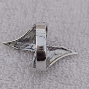 May include: A silver ring with a wing-like design. The ring has a polished finish and a detailed, textured wing shape extending from the band. The ring is set against a white, textured background.