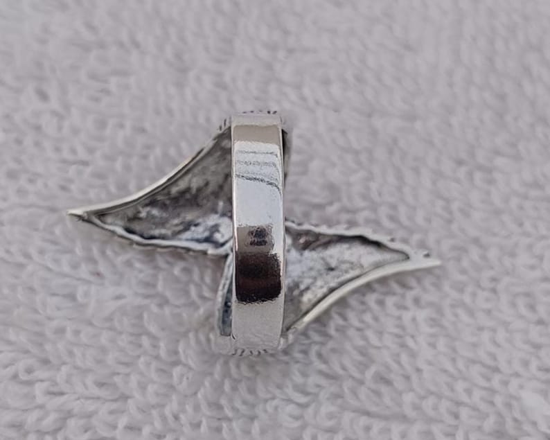 May include: A silver ring with a wing-like design. The ring has a polished finish and a detailed, textured wing shape extending from the band. The ring is set against a white, textured background.