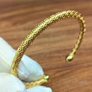 May include: A gold cuff bracelet featuring a repeating hexagonal honeycomb pattern across its entire surface. The bracelet has a rounded, open-ended design with small, smooth spherical terminals at each end, showcasing a textured metallic finish.