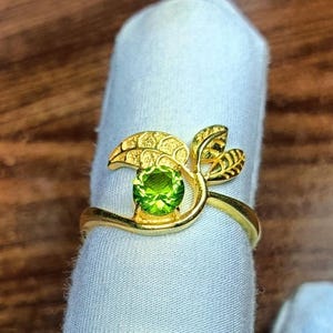 May include: A gold ring featuring a round, faceted, bright green peridot gemstone set in the centre. The band of the ring is designed with two textured, leaf-shaped accents that extend from the side of the stone, creating a botanical motif.