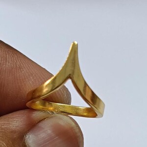 Pode incluir: Um anel de cor dourada com um design em forma de V ou chevron que se eleva até uma ponta única no topo. A banda tem um acabamento liso e polido e uma construção metálica contínua, formando uma forma geométrica simples no dedo.