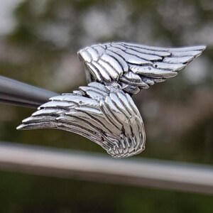 May include: A detailed, silver-colored wing pendant. The pendant features intricate feather detailing, with each feather individually defined. The wings are slightly curved, giving a sense of movement. The piece is likely jewelry, possibly a necklace charm or earring.