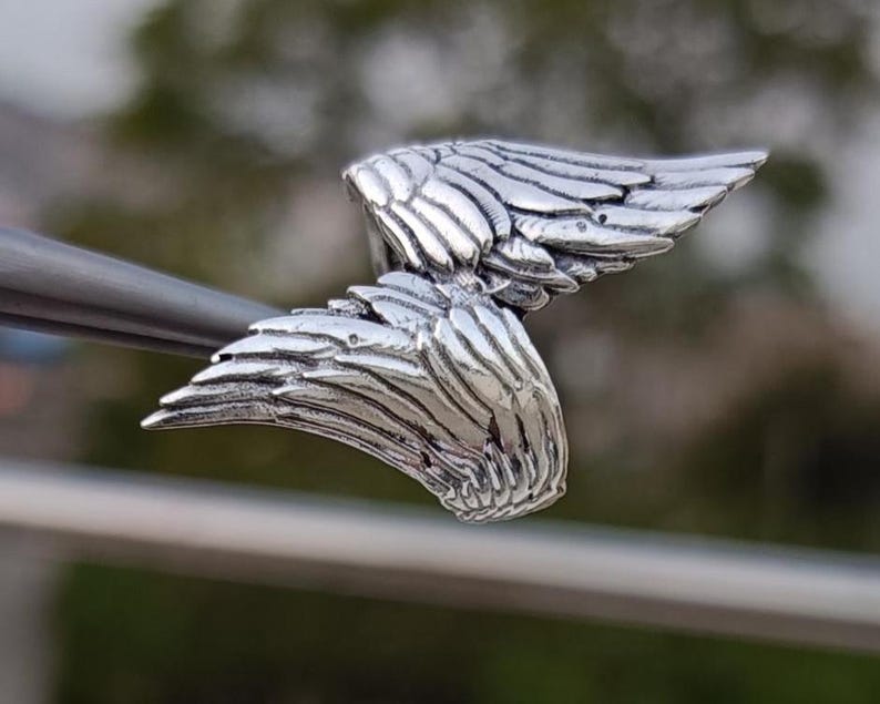 May include: A detailed, silver-colored wing pendant. The pendant features intricate feather detailing, with each feather individually defined. The wings are slightly curved, giving a sense of movement. The piece is likely jewelry, possibly a necklace charm or earring.