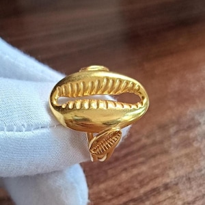 May include: A gold-toned ring featuring a prominent, horizontally oriented cowrie shell design on the face. The shell motif has a smooth, polished surface with a central slit and ribbed detailing. A smaller, matching cowrie shell charm is attached.