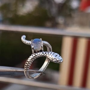 Könnte beinhalten: Ein silberner Ring in Form einer Schlange mit strukturiertem Körper und einem Edelstein. Der Schlangenkopf ist mit Schuppen und einem kleinen, runden Edelstein detailliert. Der Ring wird von einer Pinzette gehalten.