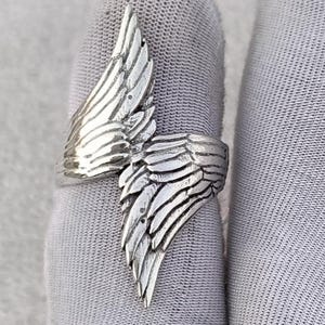 May include: A silver-colored ring with a detailed wing design. The ring features a wrap-around style with the wing extending across the finger. The wing's feathers are intricately carved, creating a textured appearance. The ring is shown on a gray surface.