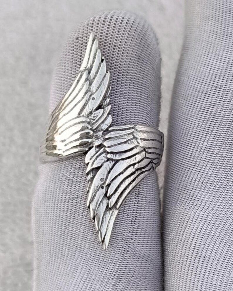 May include: A silver-colored ring with a detailed wing design. The ring features a wrap-around style with the wing extending across the finger. The wing's feathers are intricately carved, creating a textured appearance. The ring is shown on a gray surface.