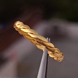 Può includere: Un anello dorato con un design a corda intrecciata. L'anello è tenuto da uno strumento argentato, che mette in risalto i suoi dettagli intricati e la finitura lucida. Il design dell'anello presenta fili intrecciati, creando un aspetto strutturato ed elegante.