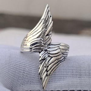 May include: A silver-colored ring with a detailed wing design. The ring features two wings that wrap around the finger, with each wing showcasing intricate feather detailing. The ring is set against a white background.
