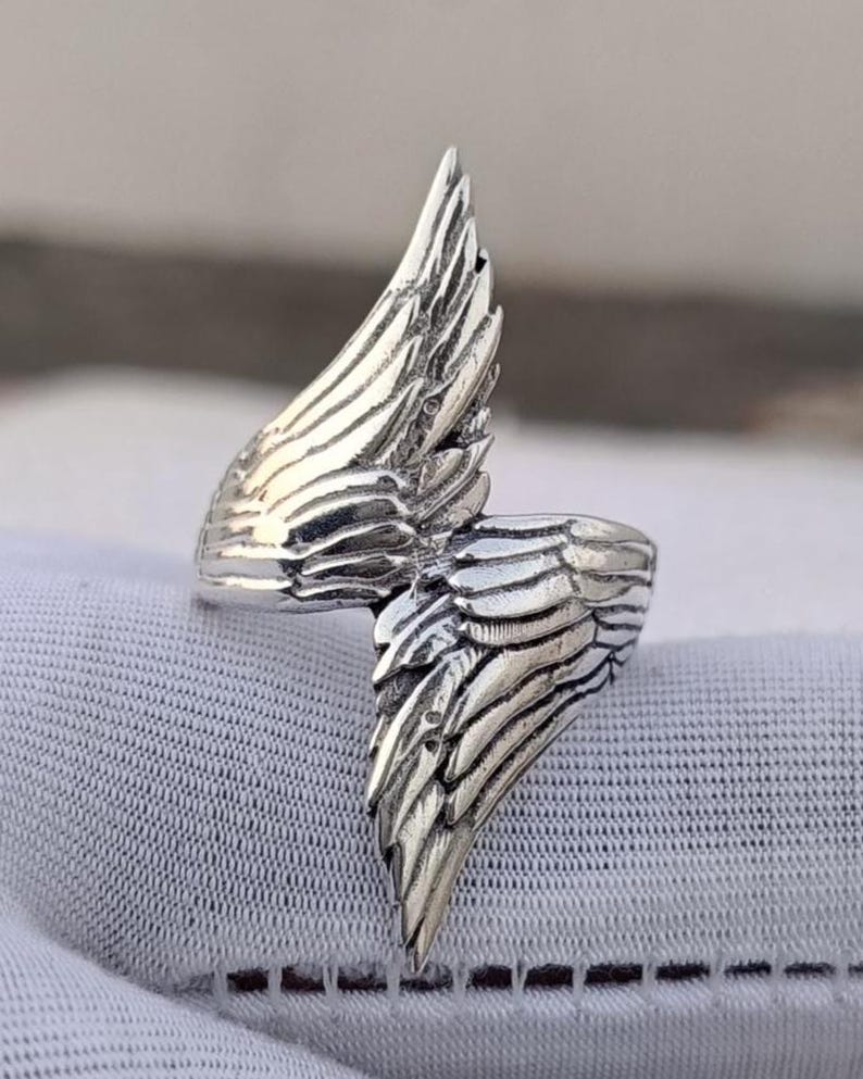 May include: A silver-colored ring with a detailed wing design. The ring features two wings that wrap around the finger, with each wing showcasing intricate feather detailing. The ring is set against a white background.