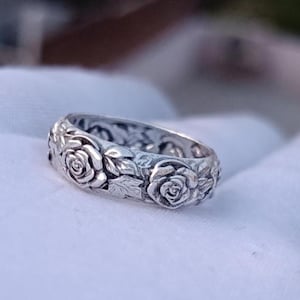 Könnte beinhalten: Ein silberner Ring mit floralem Design, das Rosen und Blätter zeigt. Der Ring hat ein detailliertes, geprägtes Muster um das gesamte Band. Der Ring wird auf einer weißen Oberfläche präsentiert.
