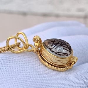 May include: A gold-colored pendant necklace with an oval-shaped gemstone. The gemstone is clear with black inclusions. The pendant has a decorative design and is attached to a delicate chain. The necklace is displayed on a white surface.