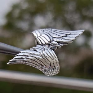 May include: A detailed, silver-colored wing pendant. The pendant features two sets of detailed wings, with each feather individually defined. The wings are slightly curved, giving a sense of movement. The pendant is held by a metal tool.