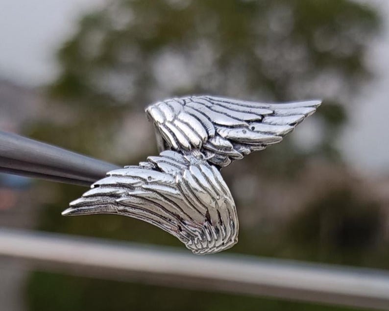 May include: A detailed, silver-colored wing pendant. The pendant features two sets of detailed wings, with each feather individually defined. The wings are slightly curved, giving a sense of movement. The pendant is held by a metal tool.