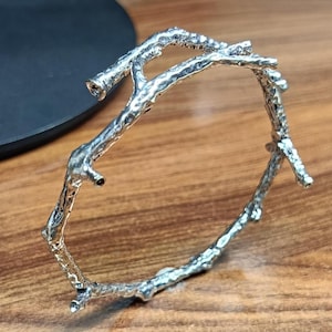 Könnte beinhalten: Ein silberfarbenes Armband in Form eines natürlichen Baumastes. Die Metalloberfläche weist eine texturierte, organische Optik mit kleinen Knoten und unregelmäßigen Erhebungen auf, die dem Aussehen einer echten Baumrinde nachempfunden ist.