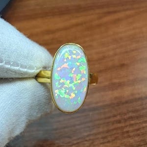 May include: A gold ring featuring a large, oval-shaped cabochon stone with a white base and iridescent, multi-colored flecks of green, orange, and blue light reflecting across the surface, set in a simple, polished gold-toned metal band.