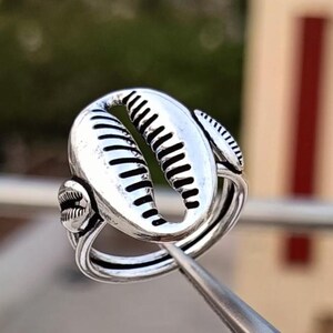 May include: A silver-colored ring featuring a seashell design. The ring has a large, oval-shaped shell in the center, with smaller shell accents on the sides. The shell details include ribbed textures and curved lines.