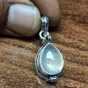 Peut inclure: Un pendentif en forme de goutte d'eau avec une pierre blanche translucide et laiteuse sertie dans une monture en argent poli. Le sertissage métallique présente des détails complexes en corde torsadée sur les bords et une bélière en argent.