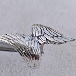 May include: A silver-colored ring featuring a detailed wing design. The ring's wings are spread out, showcasing intricate feather detailing. The ring is made of a shiny, metallic material, and the wings are the focal point of the design.