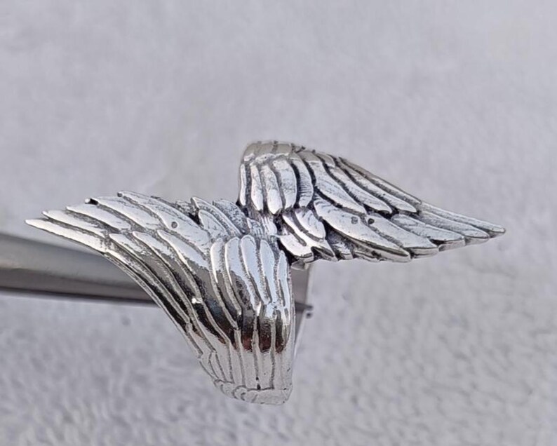 May include: A silver-colored ring featuring a detailed wing design. The ring's wings are spread out, showcasing intricate feather detailing. The ring is made of a shiny, metallic material, and the wings are the focal point of the design.