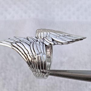 May include: A silver-colored ring with a detailed wing design. The ring features two wings extending outwards, with intricate feather detailing. The ring is held by a pair of tweezers, showcasing the craftsmanship and design.