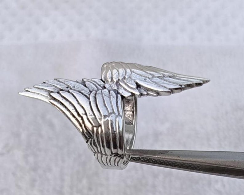 May include: A silver-colored ring with a detailed wing design. The ring features two wings extending outwards, with intricate feather detailing. The ring is held by a pair of tweezers, showcasing the craftsmanship and design.