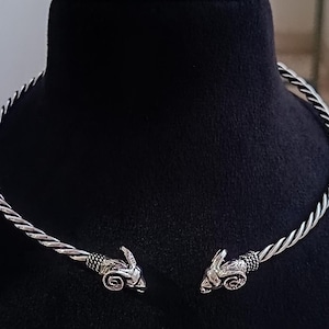 May include: A silver-toned metal torque necklace featuring a twisted rope-like band. The two open ends of the necklace terminate in detailed, sculpted ram head motifs, each showcasing curved horns and textured facial features in a polished finish.