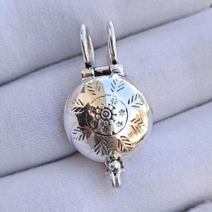 Puede incluir: Un colgante de relicario redondo de metal plateado con un motivo circular central rodeado de patrones grabados en forma de hoja. El colgante tiene una parte superior con bisagras, dos bucles verticales y un pequeño cierre.