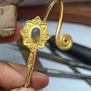Peut inclure: Un bracelet manchette en métal doré orné d'une pierre de labradorite ovale centrale sertie dans un cadre floral ouvragé. La tige présente des détails texturés près du sertissage et se termine par une boucle en spirale décorative.