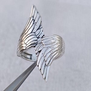 May include: A silver-colored ring featuring a detailed wing design. The ring's wings are sculpted with intricate feather details, creating a textured and dimensional appearance. The ring is shown against a neutral background, highlighting its metallic sheen.