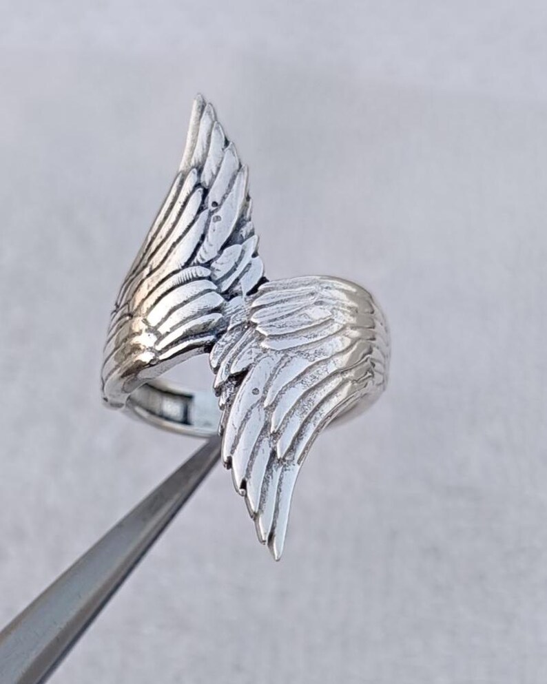 May include: A silver-colored ring featuring a detailed wing design. The ring's wings are sculpted with intricate feather details, creating a textured and dimensional appearance. The ring is shown against a neutral background, highlighting its metallic sheen.