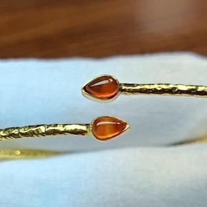 Puede incluir: Un brazalete abierto de tono dorado con una banda de metal martillado y texturizado. Los dos extremos del brazalete terminan en piedras de color naranja translúcido con forma de lágrima, creando un diseño simétrico abierto en el centro.