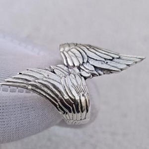 May include: A silver-colored ring featuring a detailed wing design. The wings are spread out, with intricate feather detailing. The ring appears to be made of a shiny metal, possibly sterling silver.