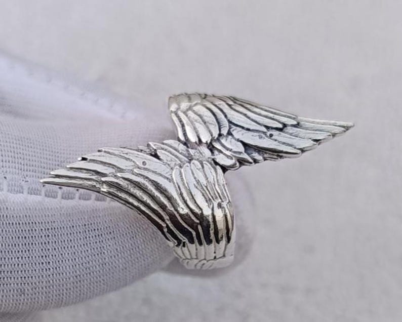 May include: A silver-colored ring featuring a detailed wing design. The wings are spread out, with intricate feather detailing. The ring appears to be made of a shiny metal, possibly sterling silver.