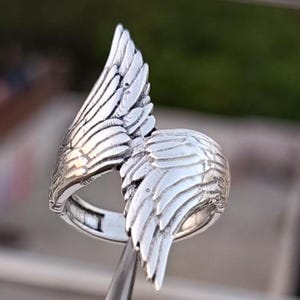 May include: A silver-colored ring with a detailed wing design. The ring features two wings, one on each side, with intricate feather detailing. The ring is held by a metal tool, showcasing the craftsmanship and design of the jewelry.
