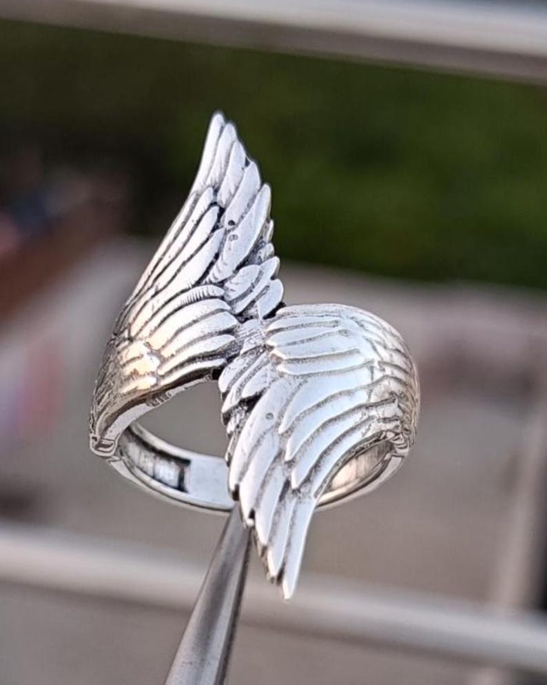 May include: A silver-colored ring with a detailed wing design. The ring features two wings, one on each side, with intricate feather detailing. The ring is held by a metal tool, showcasing the craftsmanship and design of the jewelry.