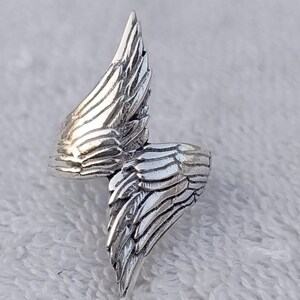 May include: A silver-colored ring with a wing design. The ring features detailed feather textures, with one wing extending upwards and the other wrapping around the finger. The ring is made of a shiny, metallic material.
