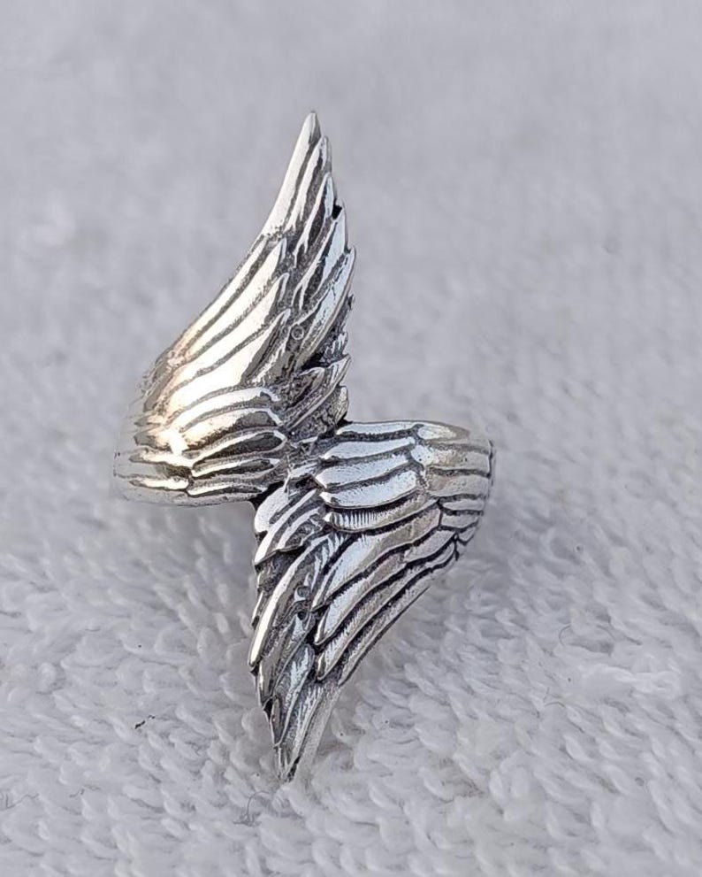 May include: A silver-colored ring with a wing design. The ring features detailed feather textures, with one wing extending upwards and the other wrapping around the finger. The ring is made of a shiny, metallic material.