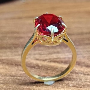 Puede incluir: Un anillo de oro con una gran piedra preciosa roja de forma ovalada y tallada, sujeta por un engaste de seis garras. La banda es lisa y pulida, mientras que la galería bajo la piedra muestra un intrincado diseño calado de inspiración floral.