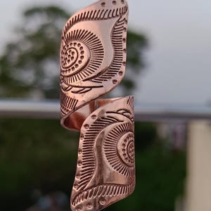 May include: Copper-colored statement ring with an open design. The ring features an intricate, engraved pattern with a central spiral and dotted details. The ring's surface has a slightly textured appearance, and the design is symmetrical.