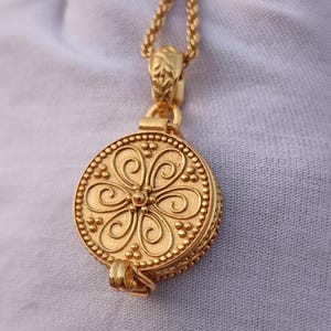 Puede incluir: Un collar con colgante dorado con un medallón redondo y ornamentado. El medallón presenta un diseño floral con detalles en espiral y un borde con cuentas. La cadena también es dorada y tiene una apariencia delicada y texturizada.
